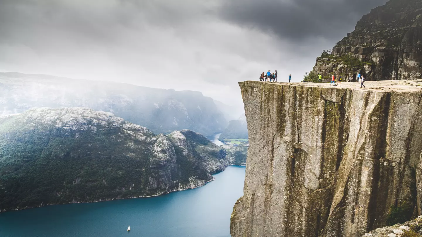 A wide shot of people standing on a the ledge atop a rocky cliff overlooking a fjord in Norway.