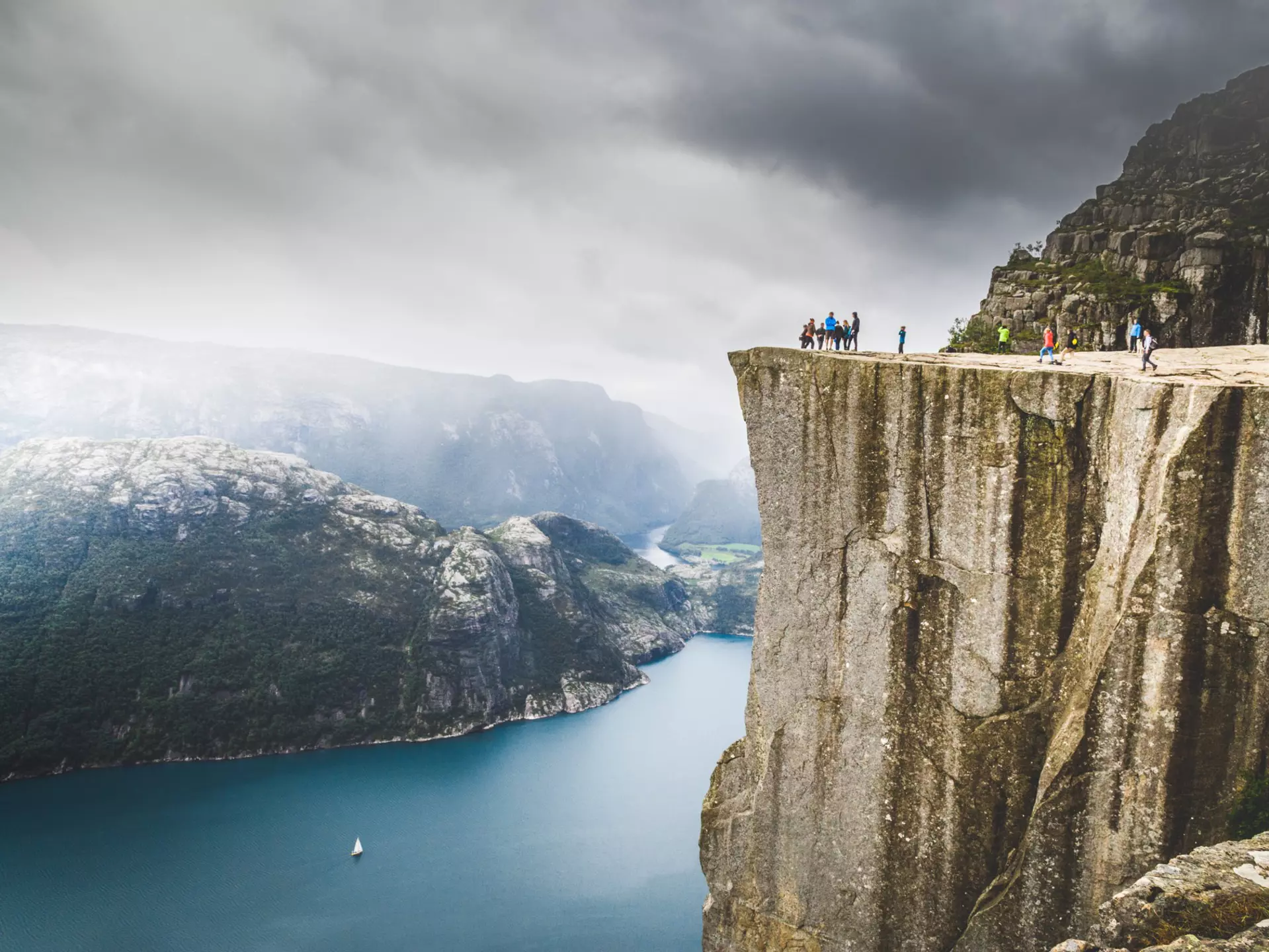 A wide shot of people standing on a the ledge atop a rocky cliff overlooking a fjord in Norway.
