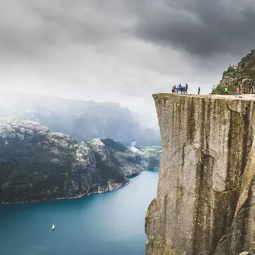A wide shot of people standing on a the ledge atop a rocky cliff overlooking a fjord in Norway.