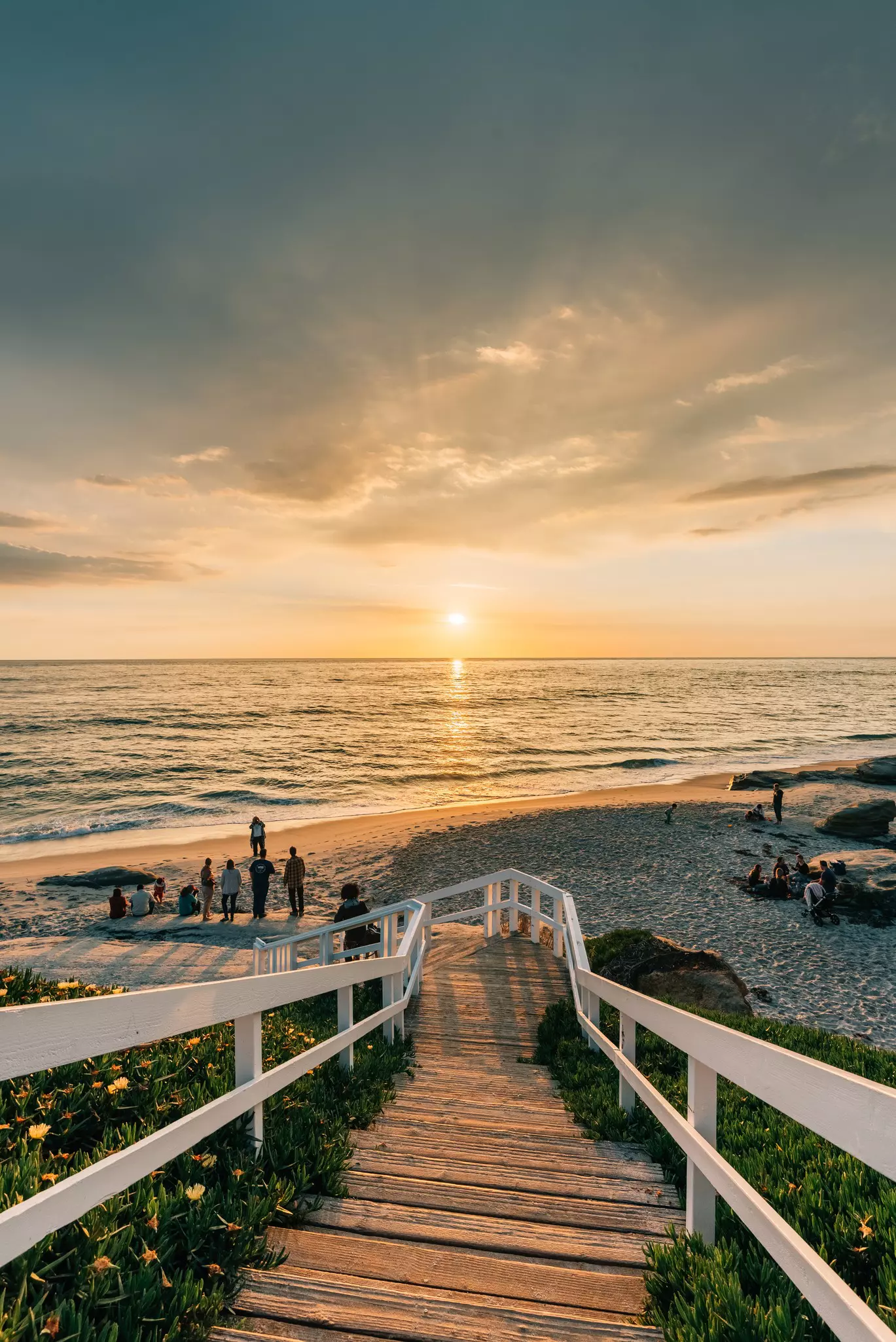A boardwalk and stairs lead down to a beach where people are watching the sunset over the sea.