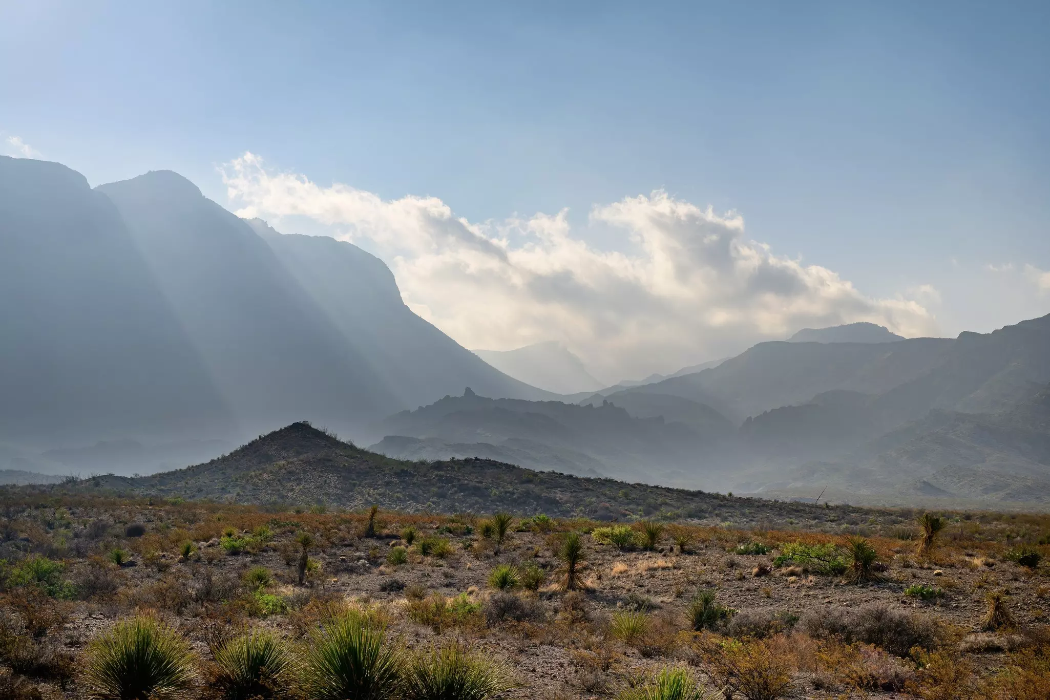 Beams of light dramatically illuminate a rocky ridge, with scrubland seen in the foreground.