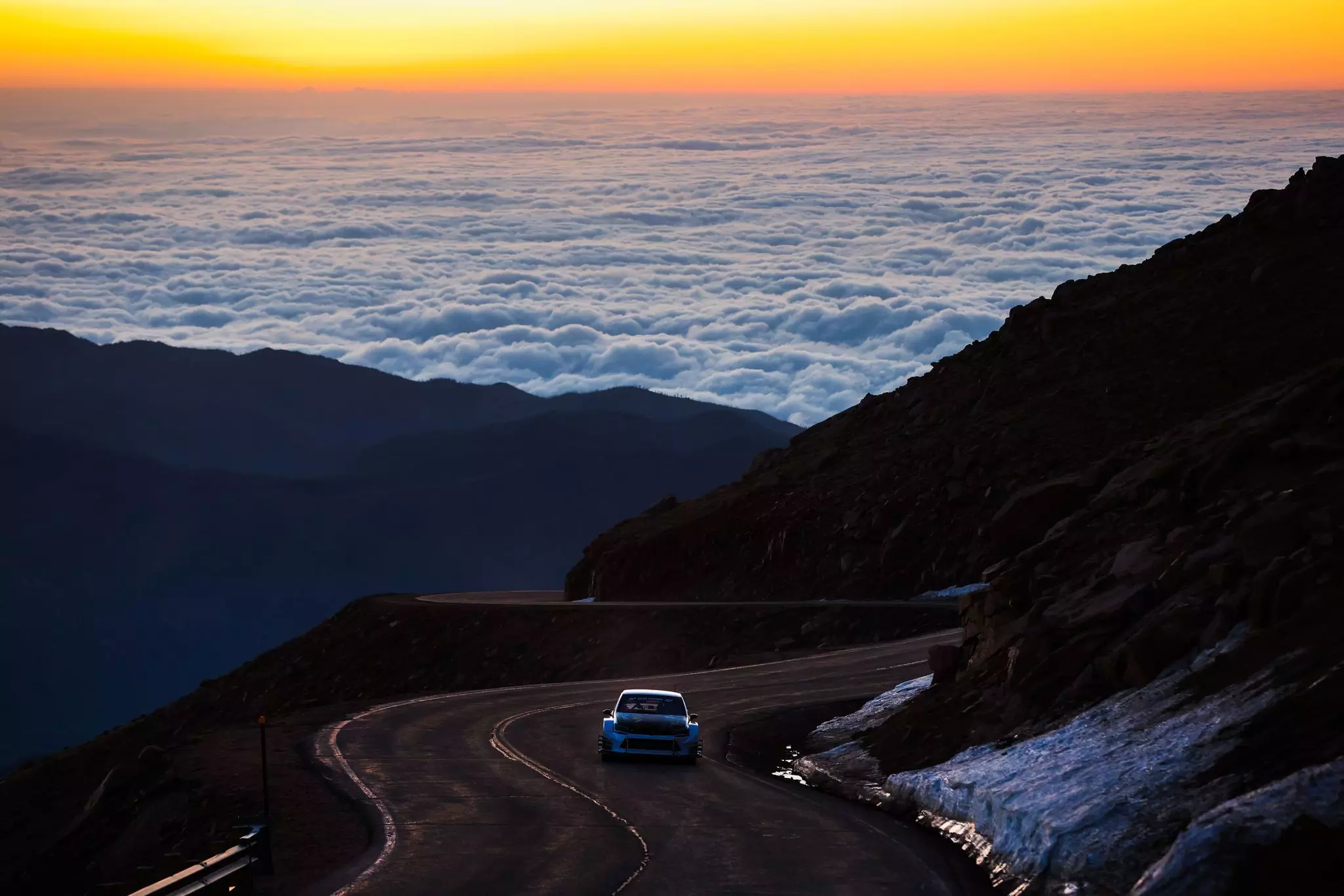 A car is seen from behind driving on the Pikes Peak Hwy above the clouds, with a line of yellow setting sun in the background
