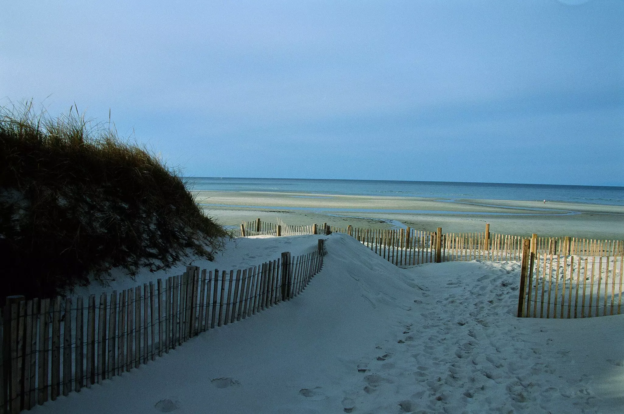 Sand fence entering through low dunes leading to a beach and the ocean beyond on a sunny day.