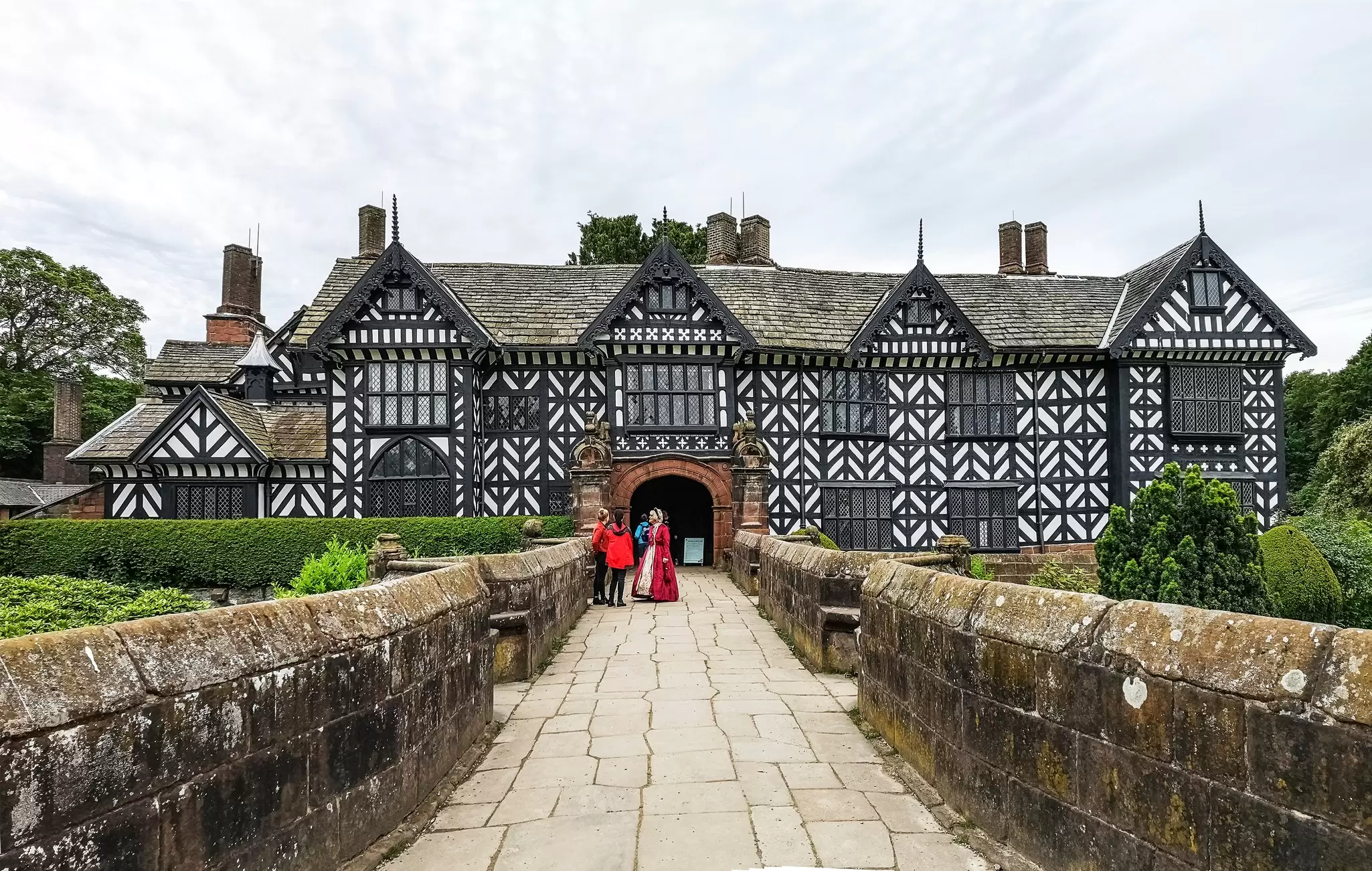 A woman in Tudor dress stands on a walkway talking to tourists visiting a huge Tudor mansion with a black and white facade.