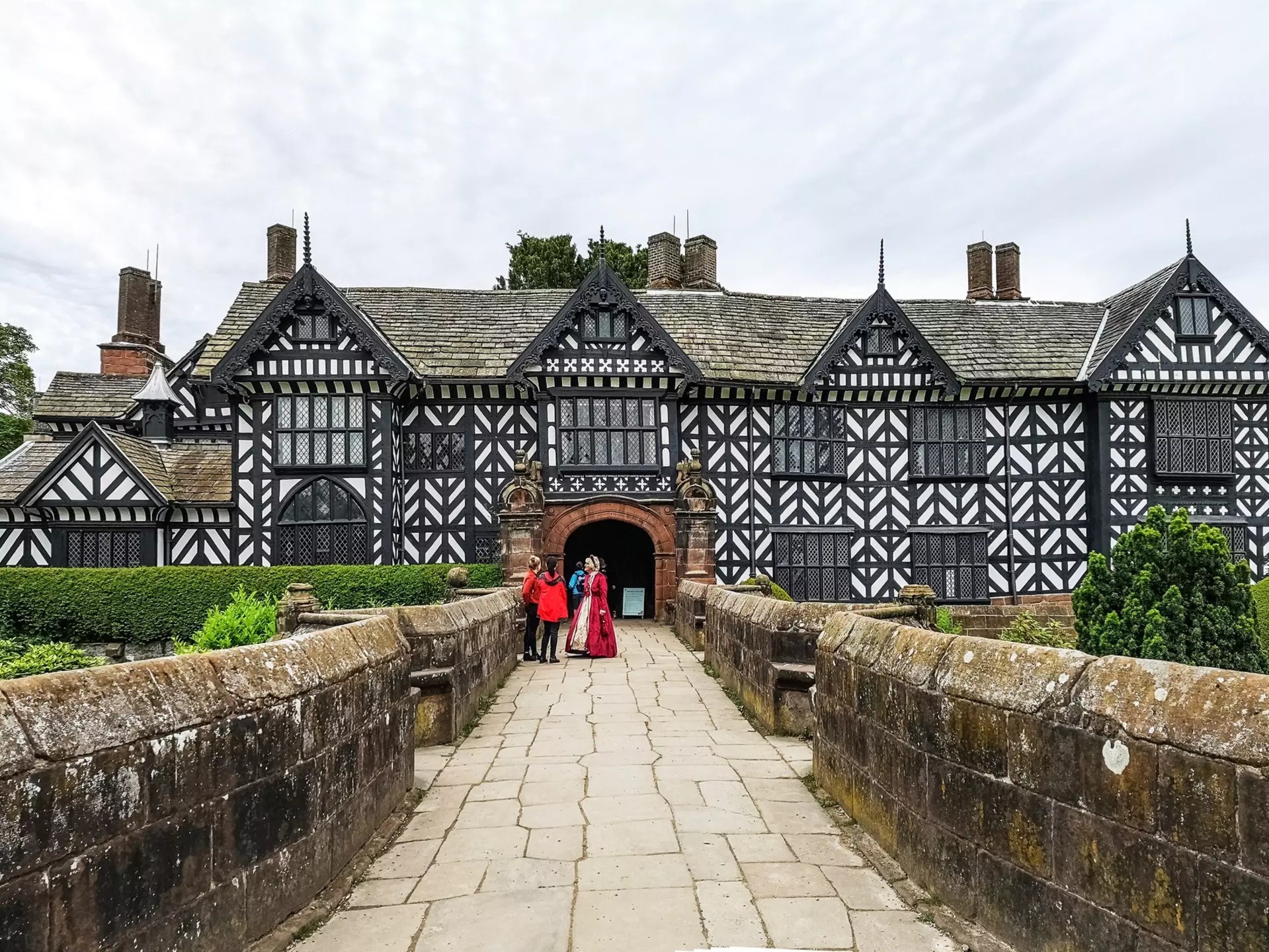 A woman in Tudor dress stands on a walkway talking to tourists visiting a huge Tudor mansion with a black and white facade.