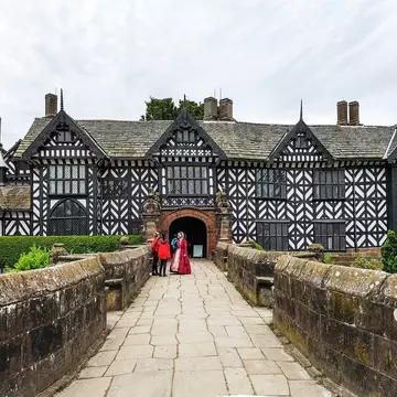 A woman in Tudor dress stands on a walkway talking to tourists visiting a huge Tudor mansion with a black and white facade.