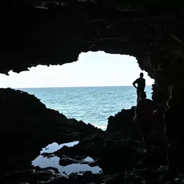 Silhouetted people stand inside a dark sea cave whose wide opening frames turquoise ocean, with rock pools on the floor.
