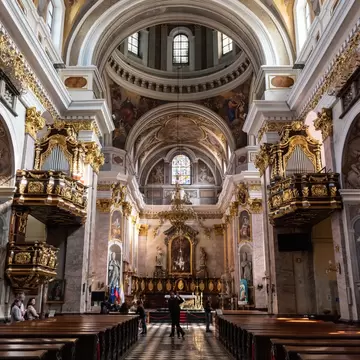 A baroque church interior with frescoed vaults, gilded organ balconies on both sides and a chequered marble floor running to the high altar.