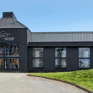 A dark grey metal-clad distillery building with tall windows and a pitched roof, signed Isle of Raasay Distillery, beside a gravel drive.