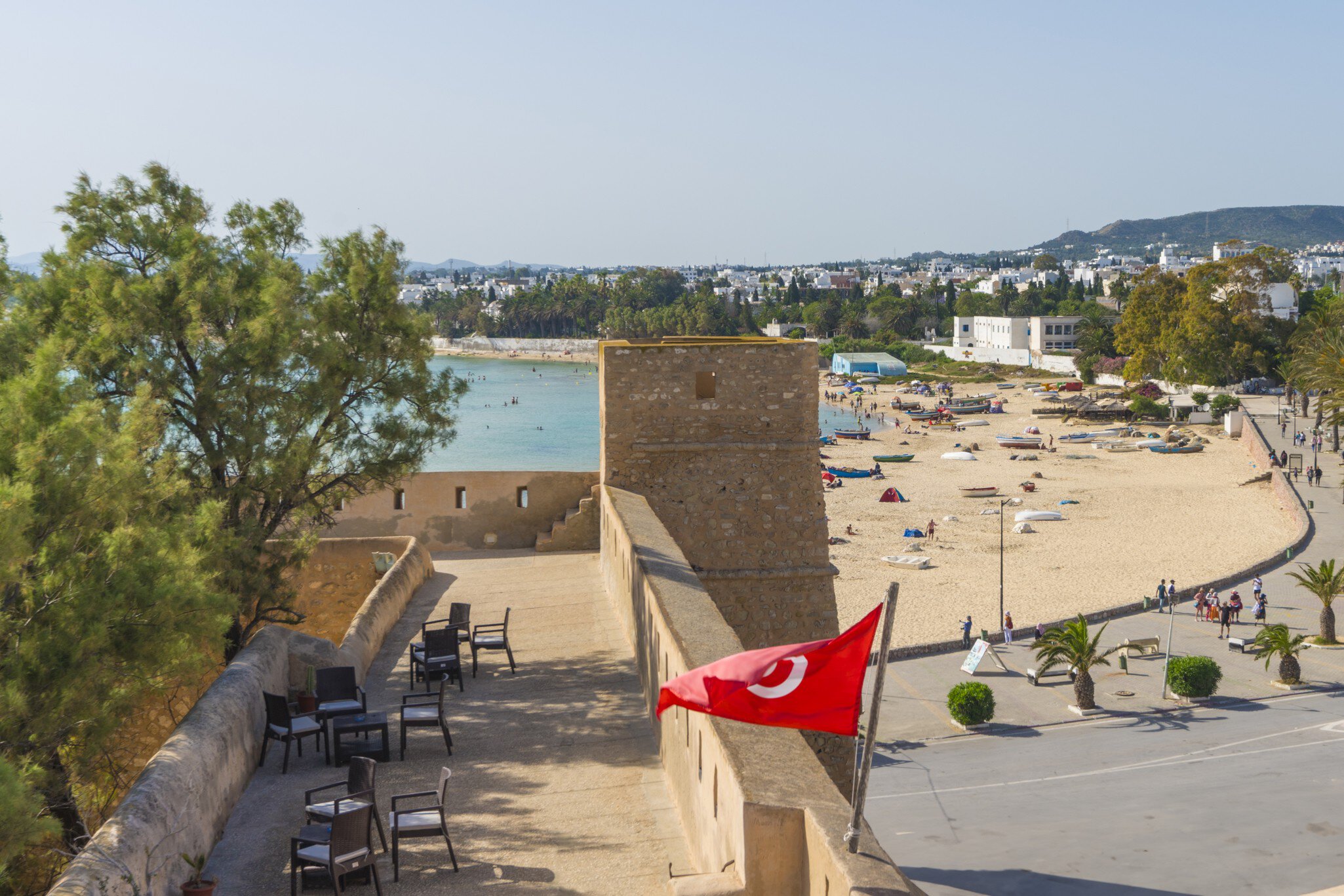 Stone ramparts with a Tunisian flag above a sandy beach lined with small boats and white buildings in Hammamet.