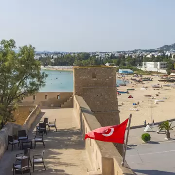 Stone ramparts with a Tunisian flag above a sandy beach lined with small boats and white buildings in Hammamet.