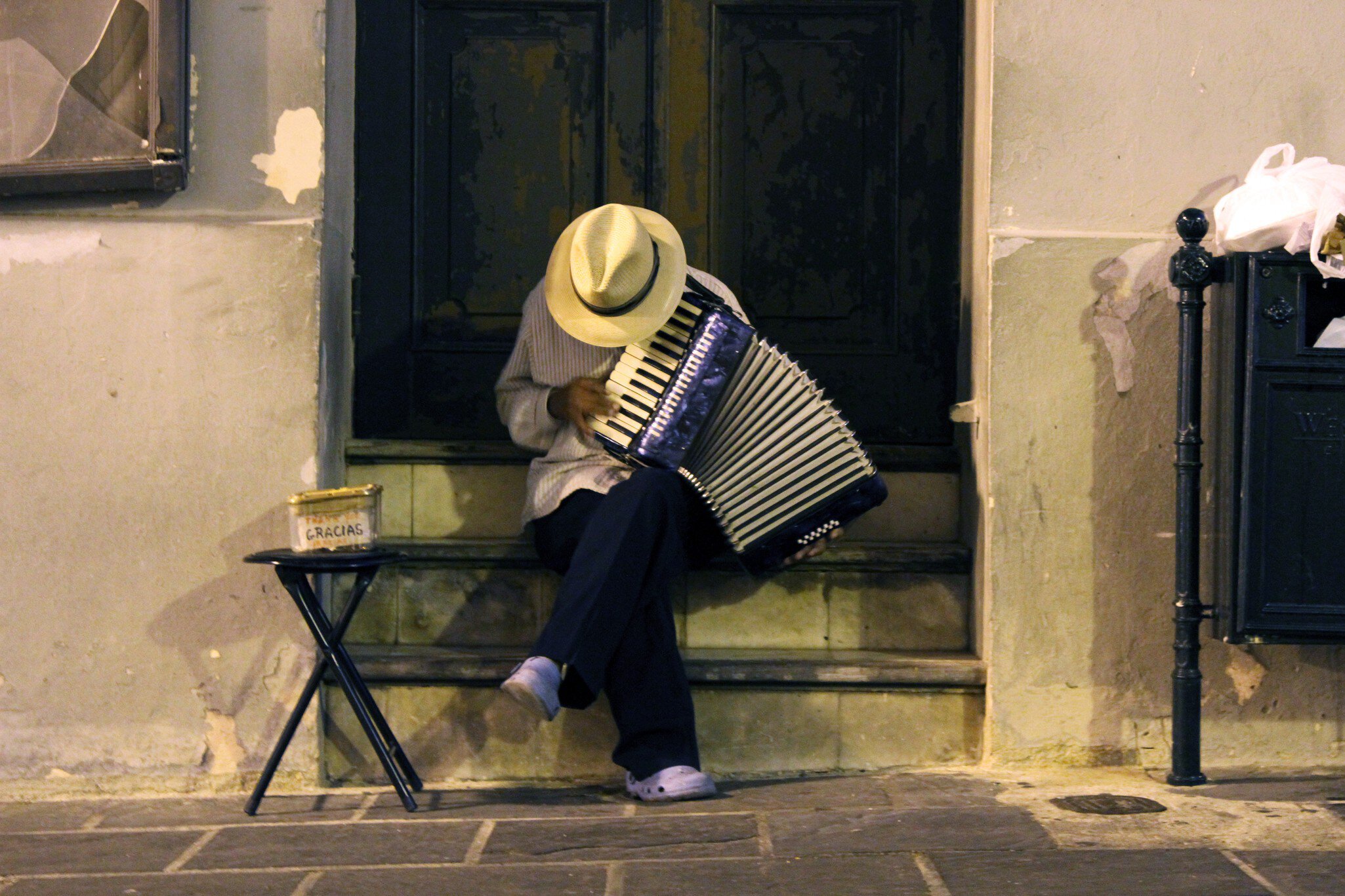 A man in a straw hat sits on a stoop, head down, playing an accordion. A small table to his right holds a metal tin with the word