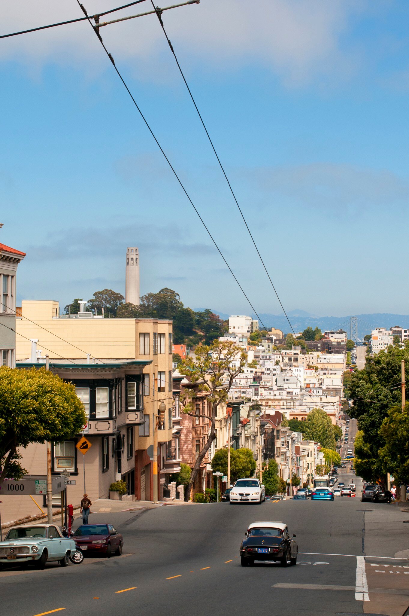 A steep street lined with pastel houses and parked cars leads toward North Beach, with Coit Tower on a wooded hill beyond.