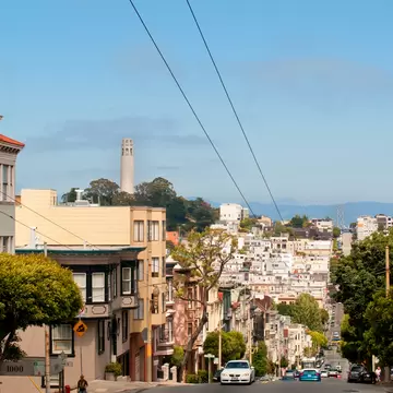 A steep street lined with pastel houses and parked cars leads toward North Beach, with Coit Tower on a wooded hill beyond.