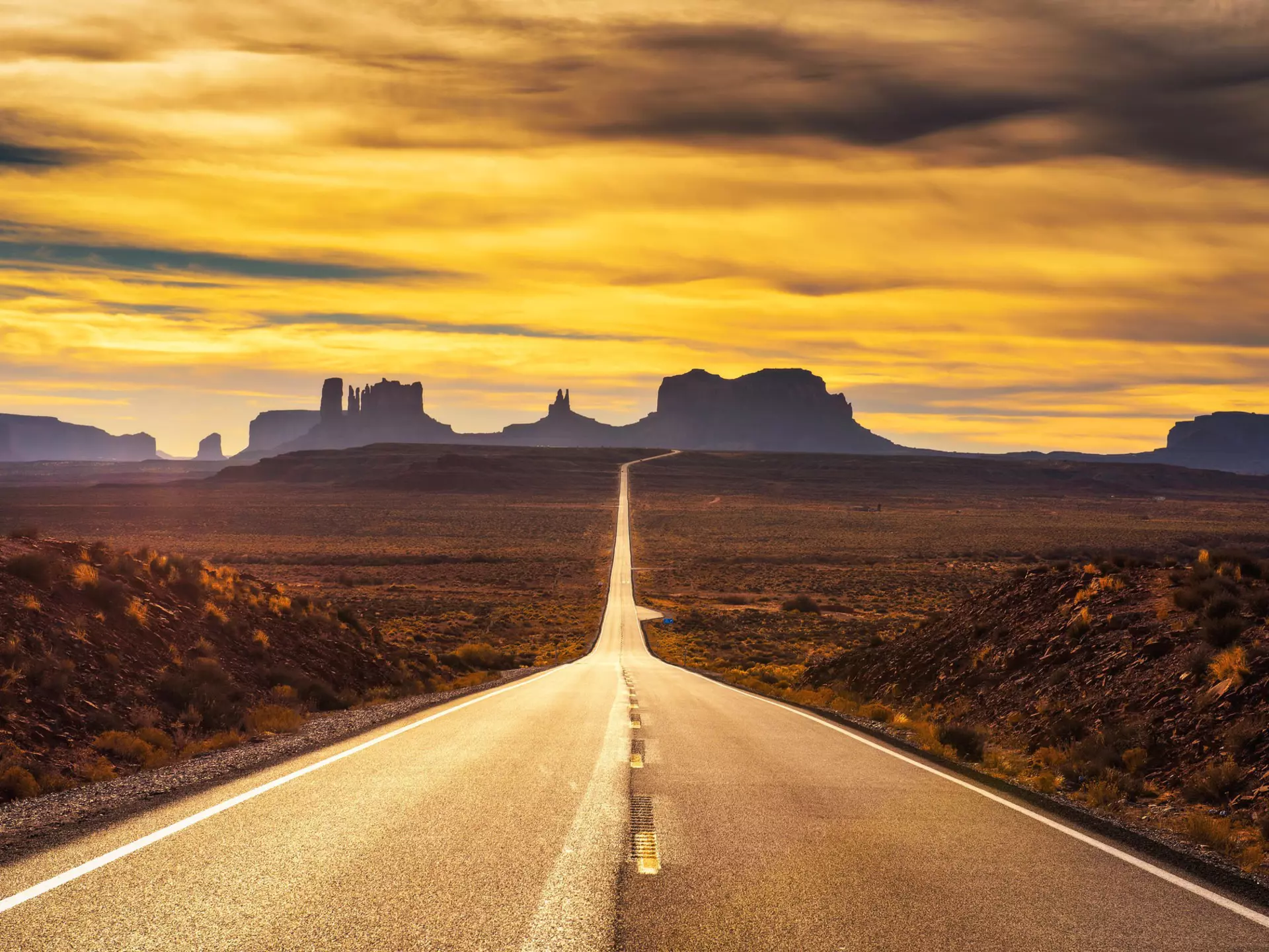 A straight strech of an empty highway leading through the desert at dusk, with the mesas and rock formations silhouetted in the distance, Arizona