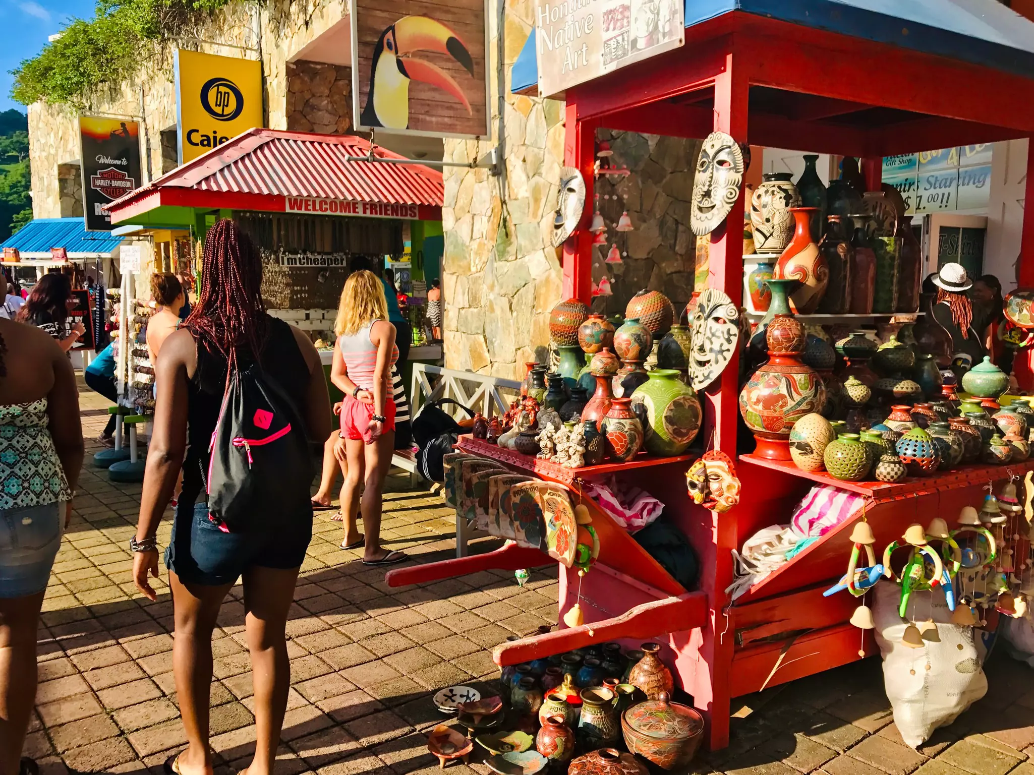 People past a red cart filled with pottery and masks for sale on a cobbled street.