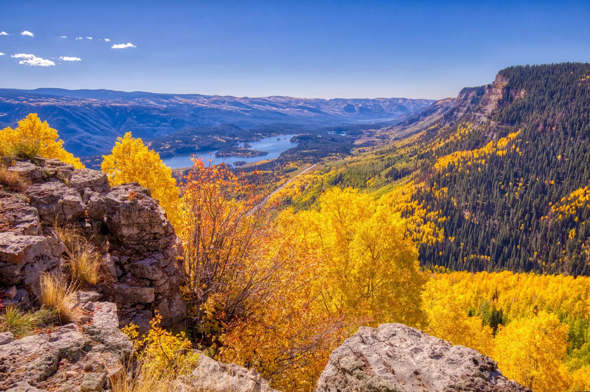 A panorama of a valley with a distant lake on a sunny day; bright yellow trees and a rock face are in the foreground.