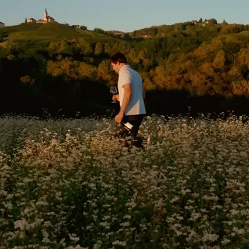 A person walks through a flowering field holding a wine glass.