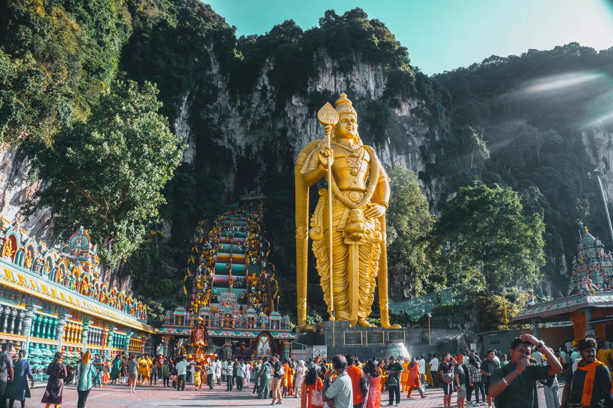 A large golden statue stands in a courtyard against a cliff in Malaysia; a crowd of visitors are in the courtyard.