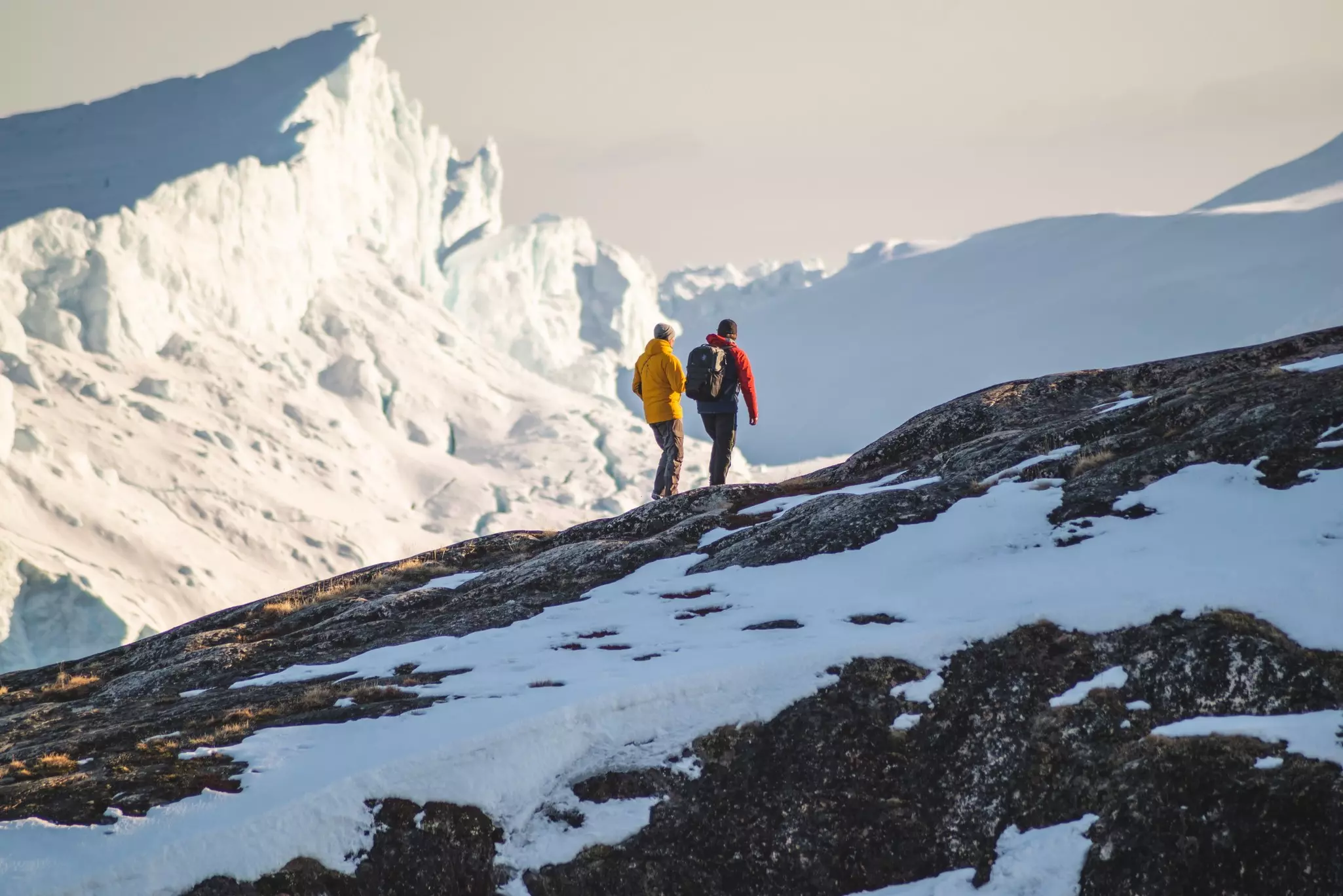 Two hikers walk up a steep rocky incline with patches of snow; a wedge of a white glacier is in the background.