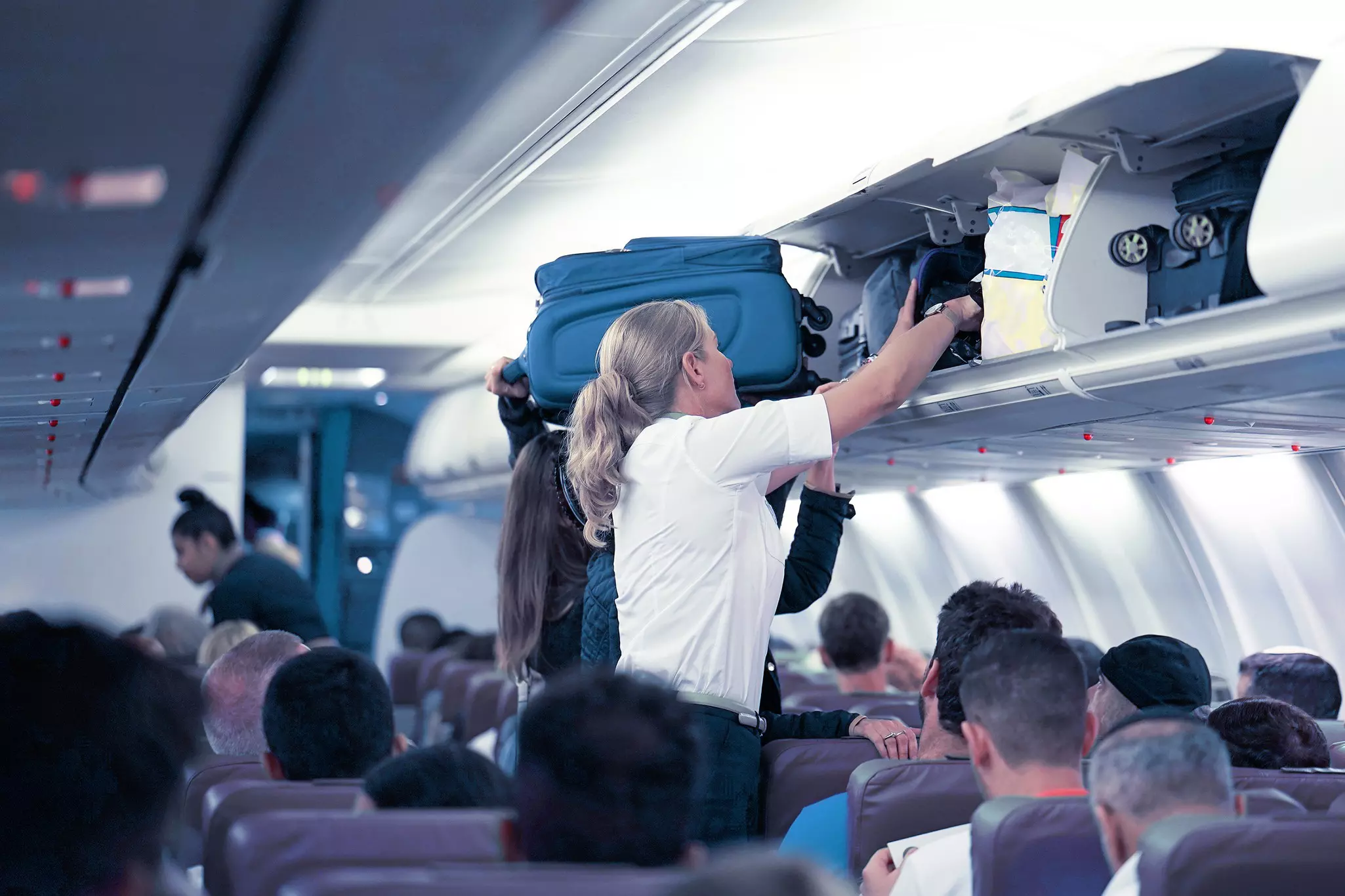 An air steward and passenger load bags into the overhead lockers on a plane before departure.