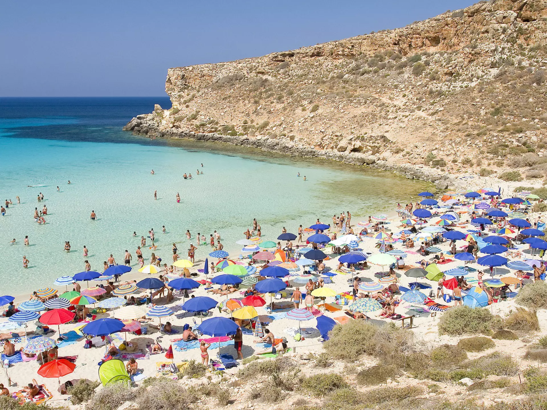 Tourists on a busy beach taking shade under umbrellas and swimming in the shallow calm waters.