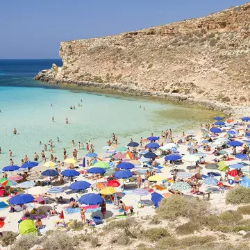 Tourists on a busy beach taking shade under umbrellas and swimming in the shallow calm waters.