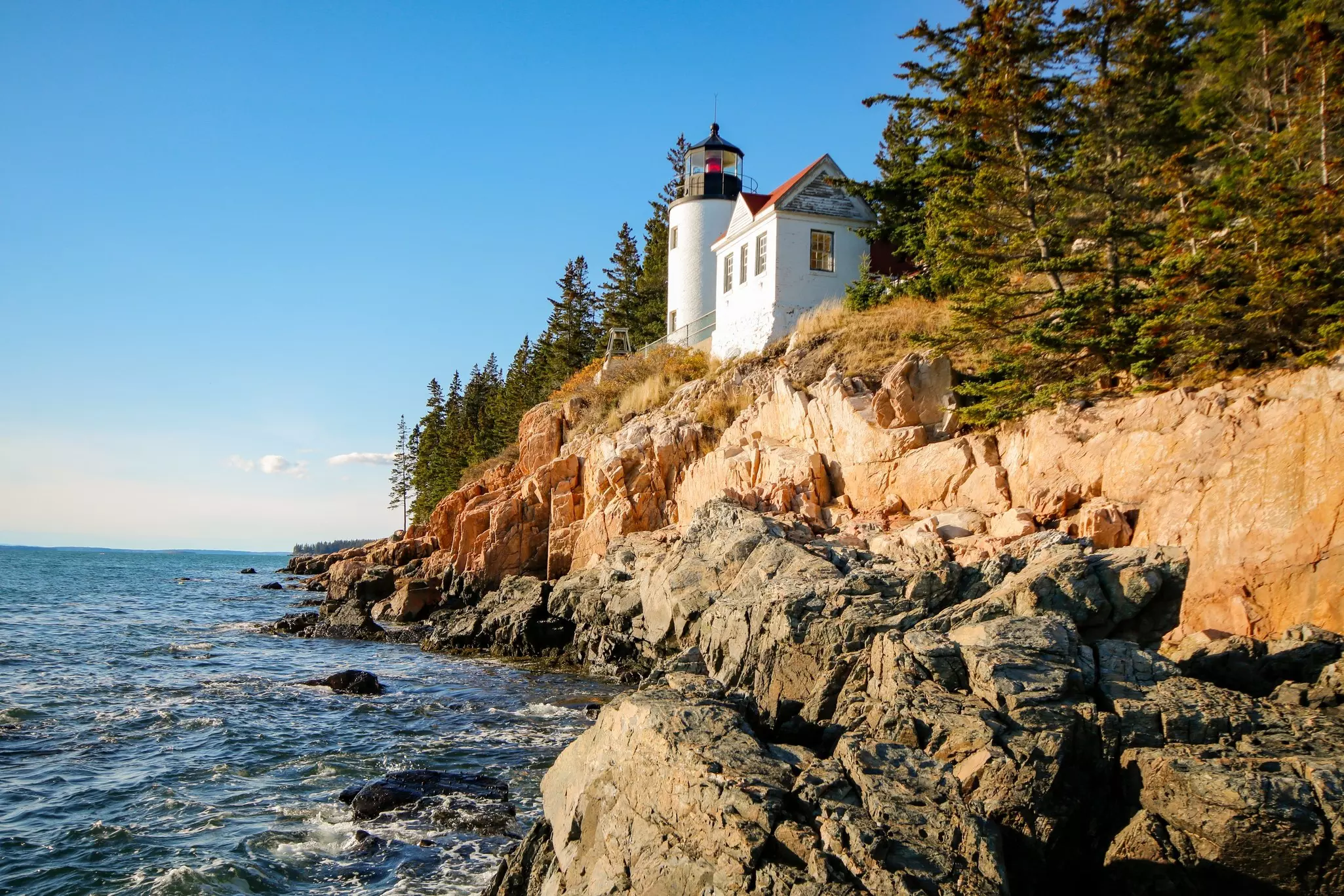 White building and lighthouse on a rocky cliff above dark blue ocean on a bright, sunny day.