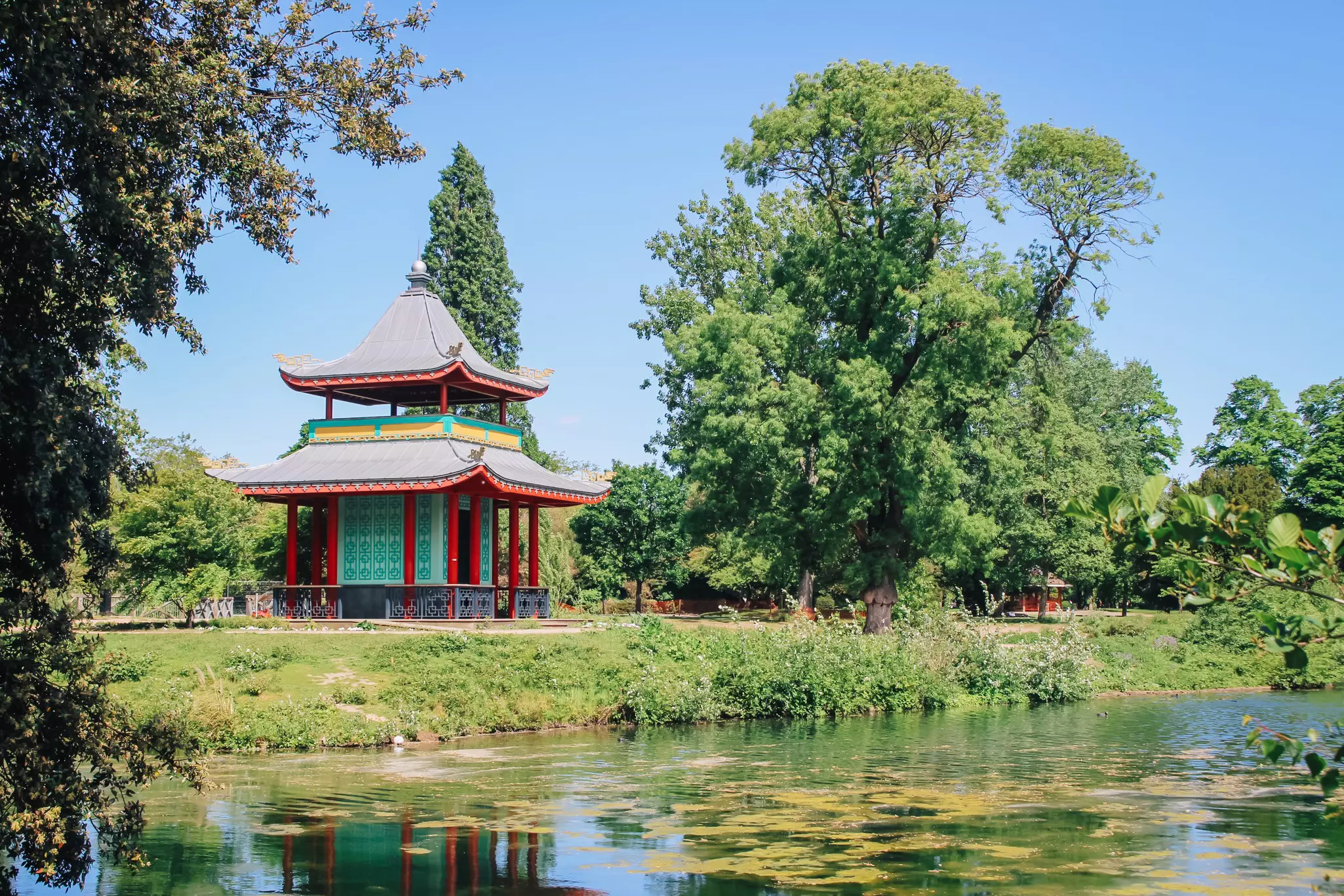 Chinese pagoda on a grassy bank near a pond and surrounded by trees on a sunny day.