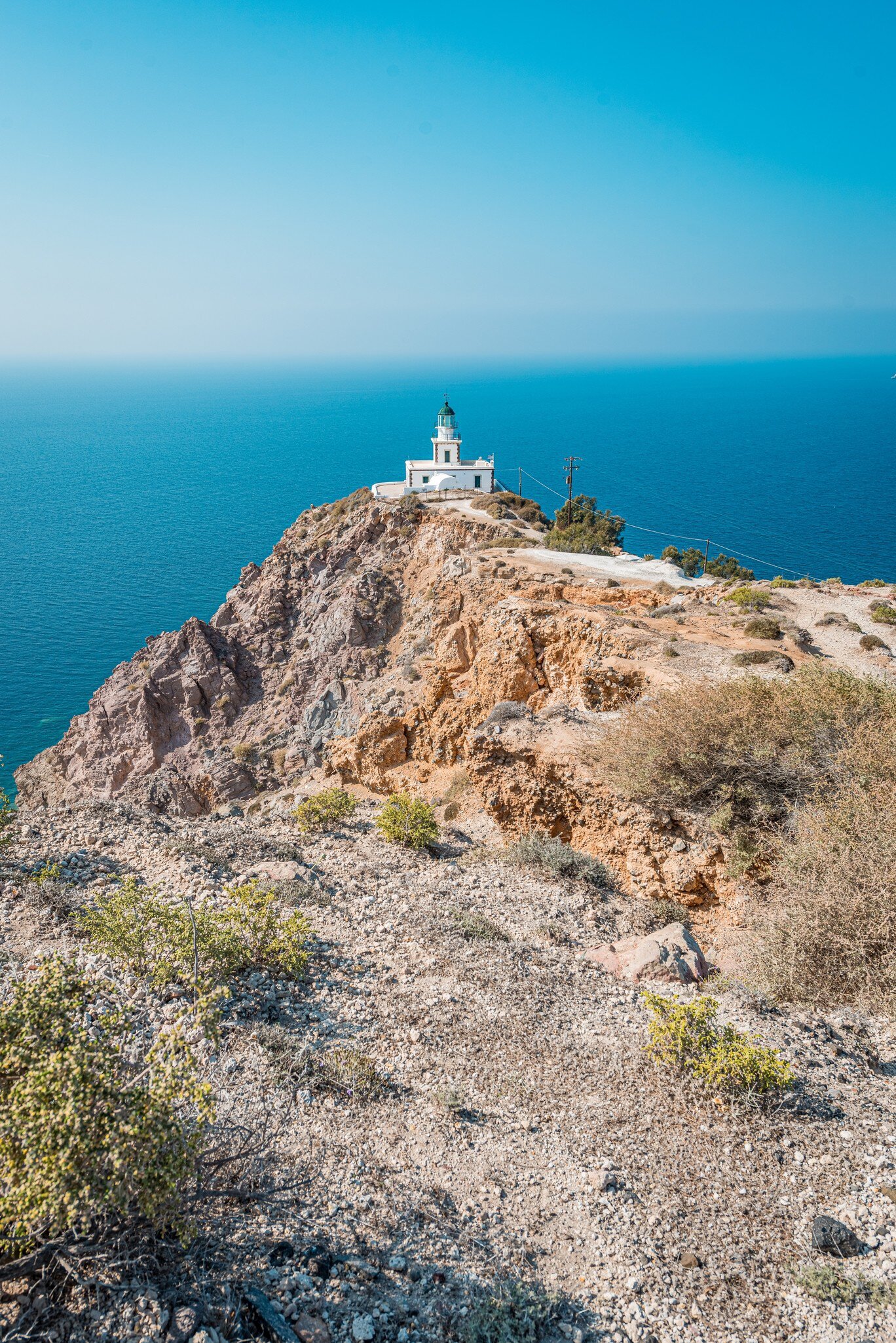 Akrotiri Lighthouse, a 19th-century lighthouse on the Greek island of Santorini with sun reflecting off of the clear blue water in the background.