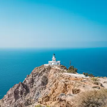 Akrotiri Lighthouse, a 19th-century lighthouse on the Greek island of Santorini with sun reflecting off of the clear blue water in the background.