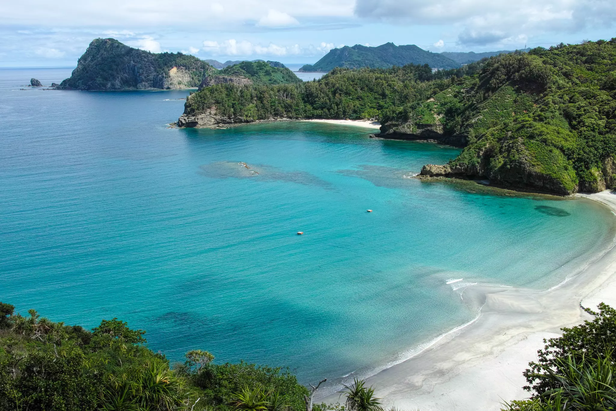 Aerial view of beach with clear blue ocean and rocky, forested islands in the distance on a sunny day.