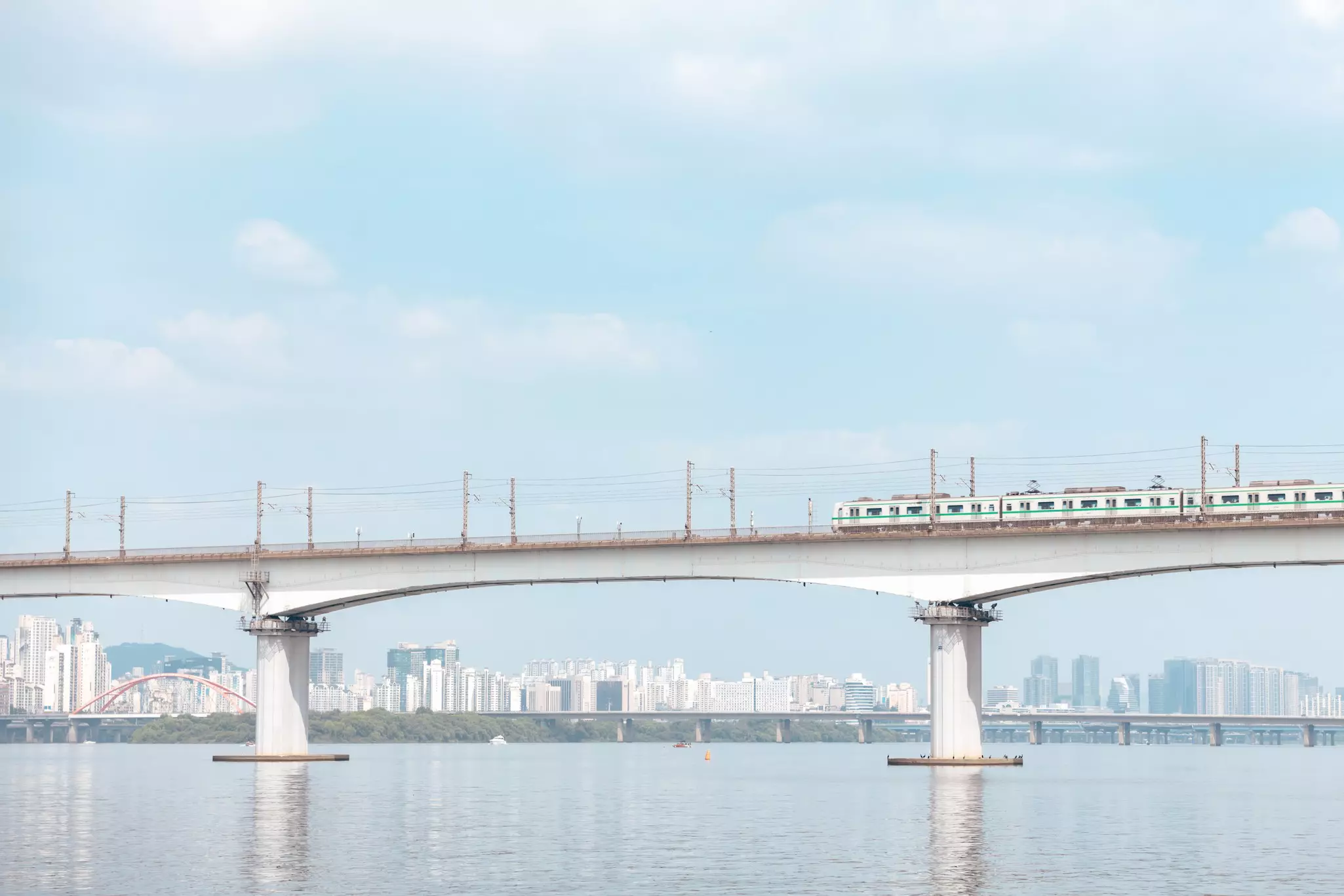 A train is seen on a bridge that crosses a wide river on a bright day. Towers of a city are seen on the shore in the distance.