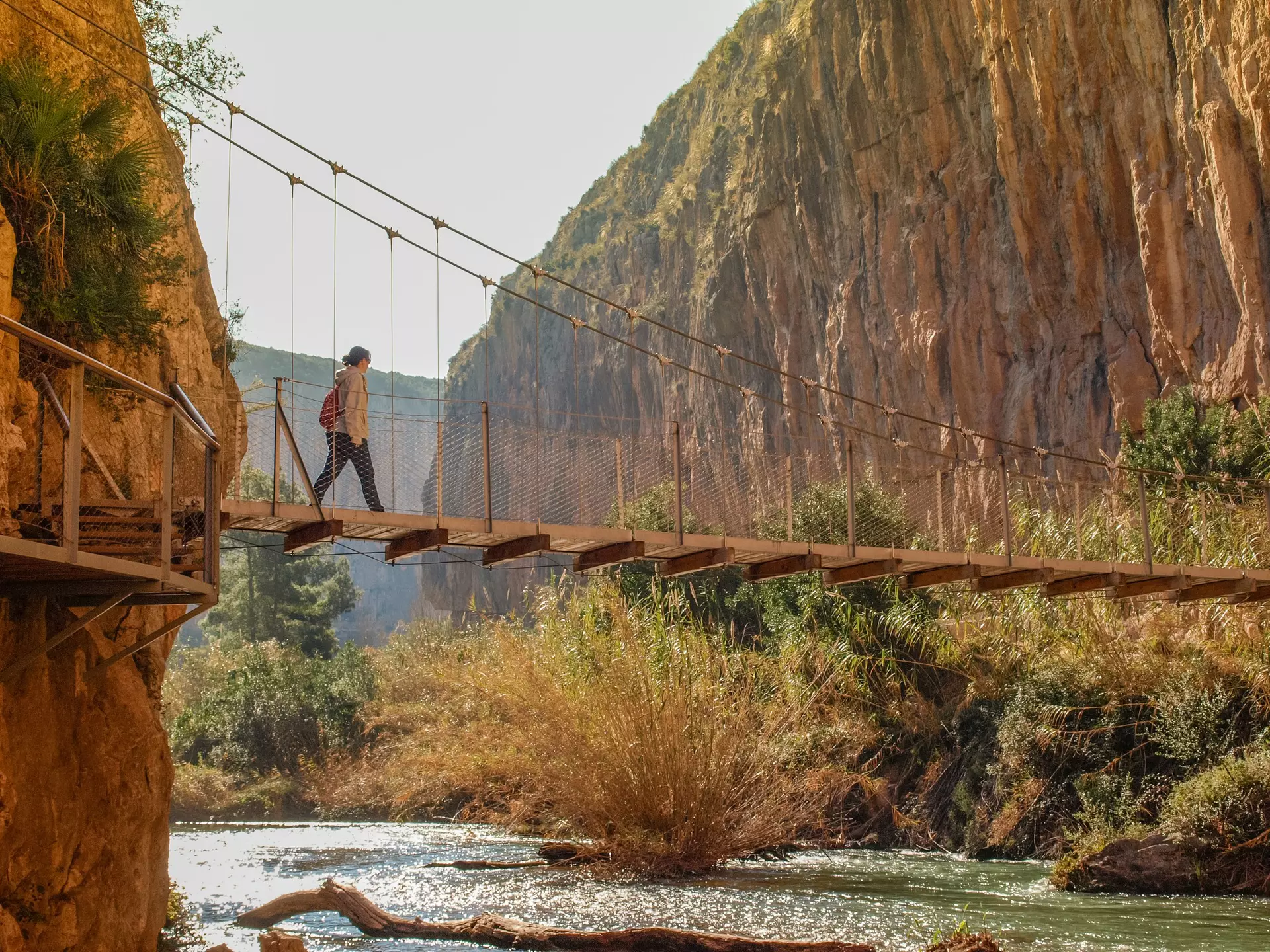 A hiker walks over a suspension bridge above a river, linking up two sides of a gorge.