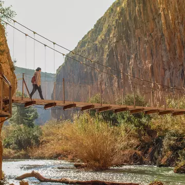 A hiker walks over a suspension bridge above a river, linking up two sides of a gorge.