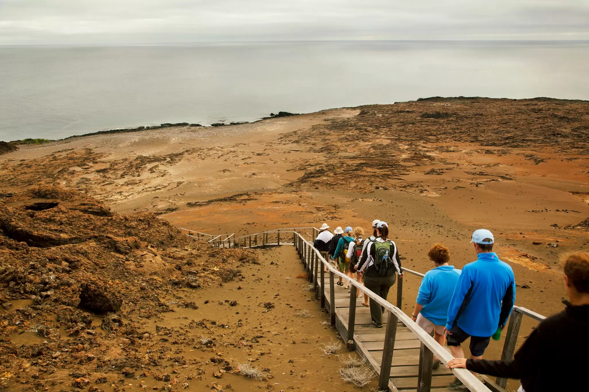 People walk on a narrow boardwalk down a barren slope of an island, the Galápagos Islands