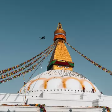 Gold tower with painted eyes above a white dome streaked with saffron, prayer flags strung across blue sky.
