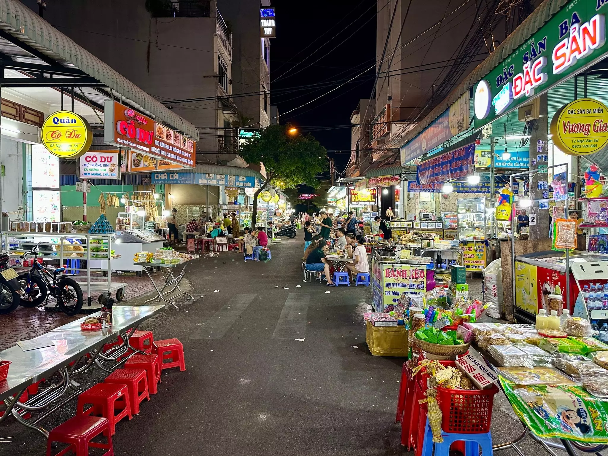 A city street in the evening lined with stalls selling food. Groups sit at small plastic stools to eat.
