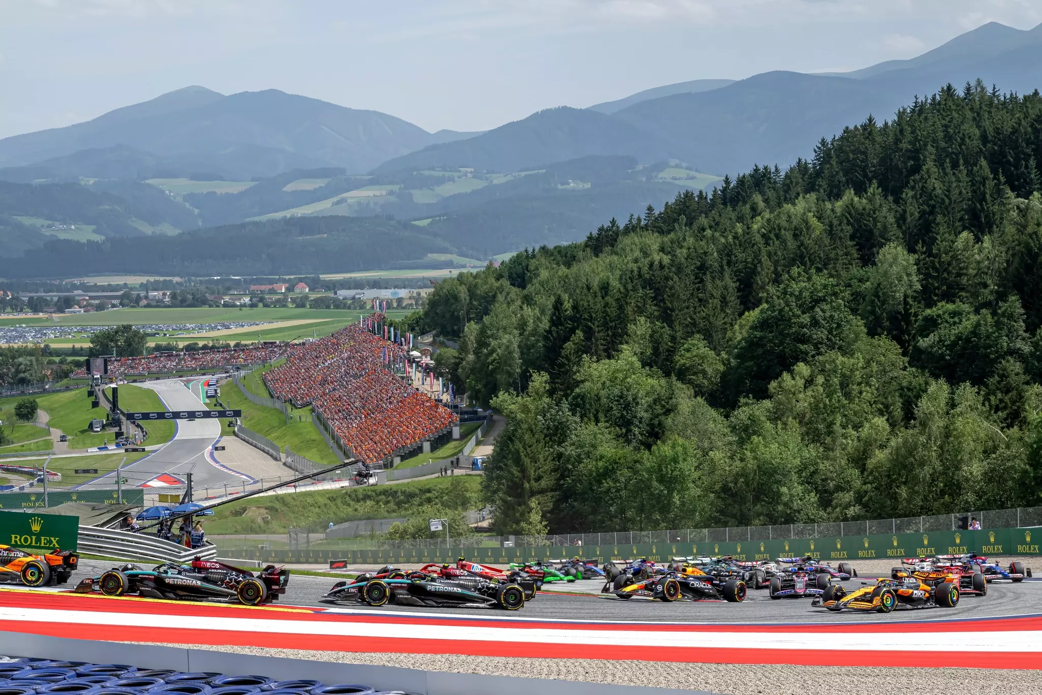 Several race cars drive on a track in a bucolic mountain setting.