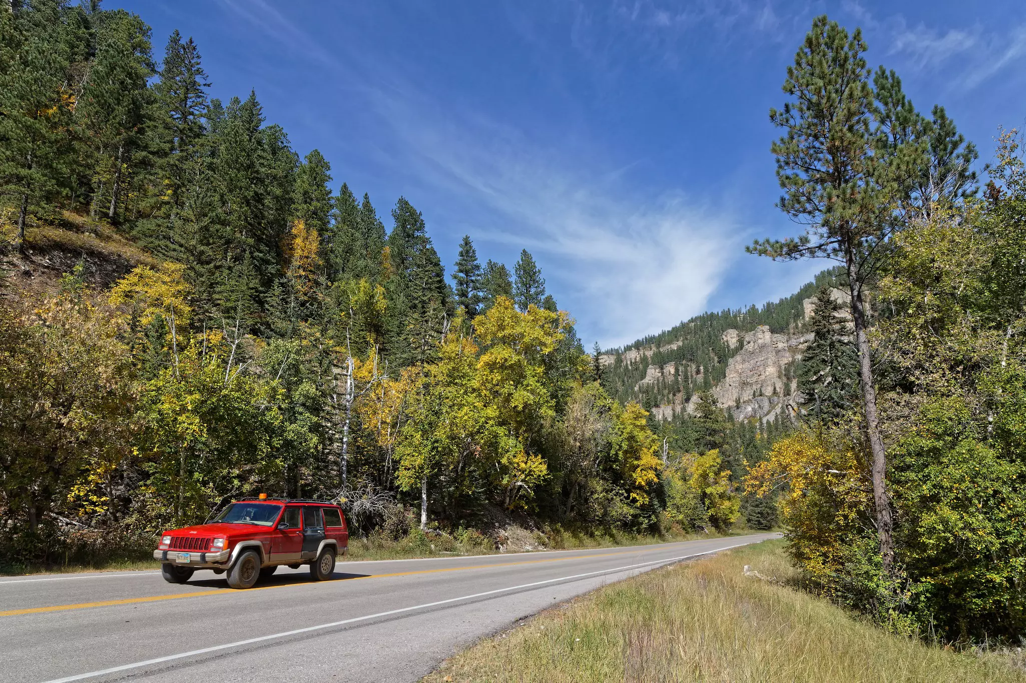 A red car drives along a road in early fall as the leaves on the trees are starting to turn from green to yellow.