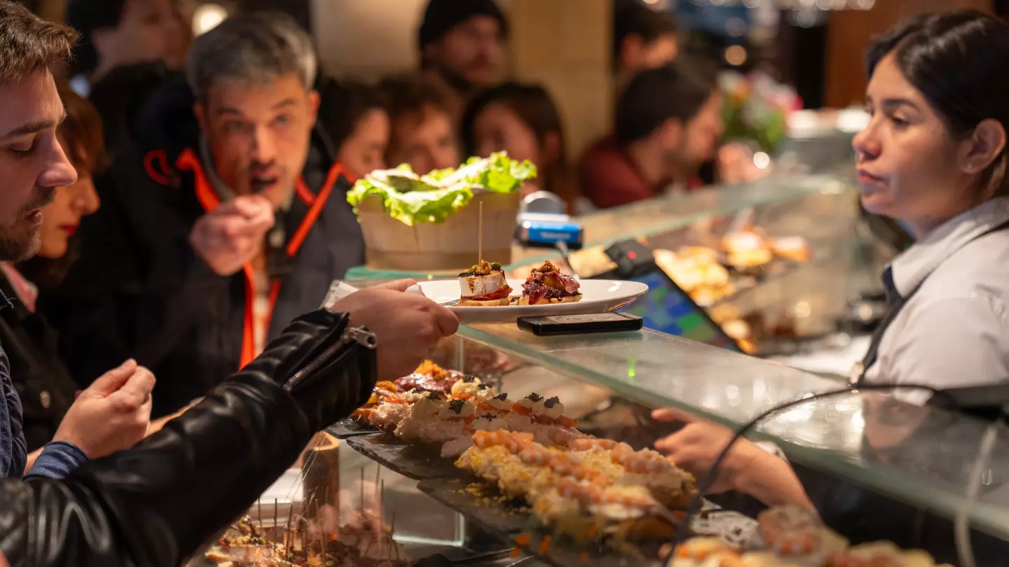 People at a bar with food displayed on shelves. One patron takes a plate from a glass shelf, with a server behind the counter.