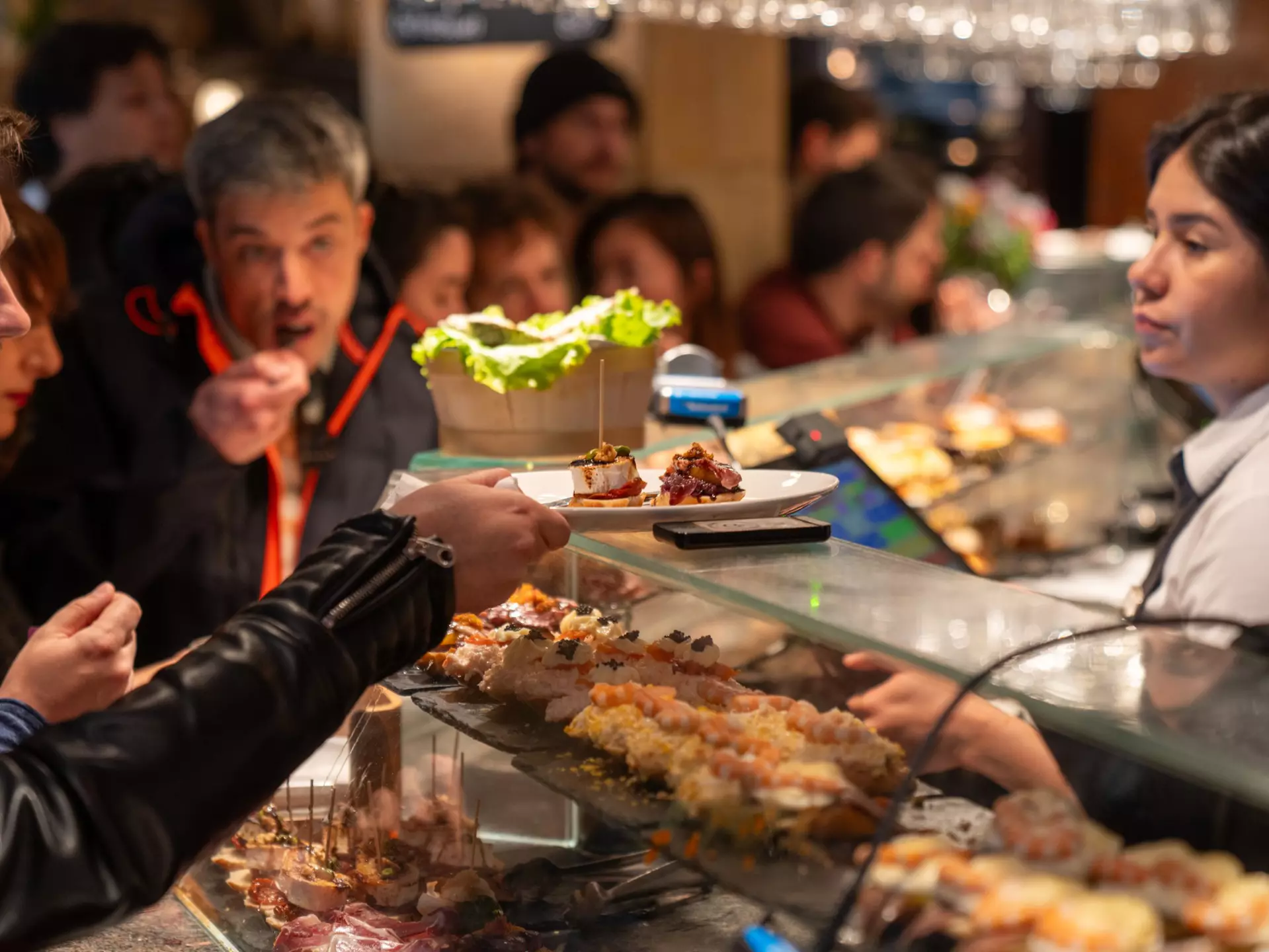 People at a bar with food displayed on shelves. One patron takes a plate from a glass shelf, with a server behind the counter.