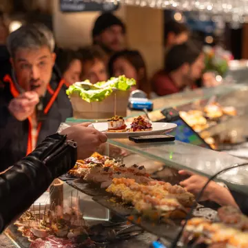 People at a bar with food displayed on shelves. One patron takes a plate from a glass shelf, with a server behind the counter.