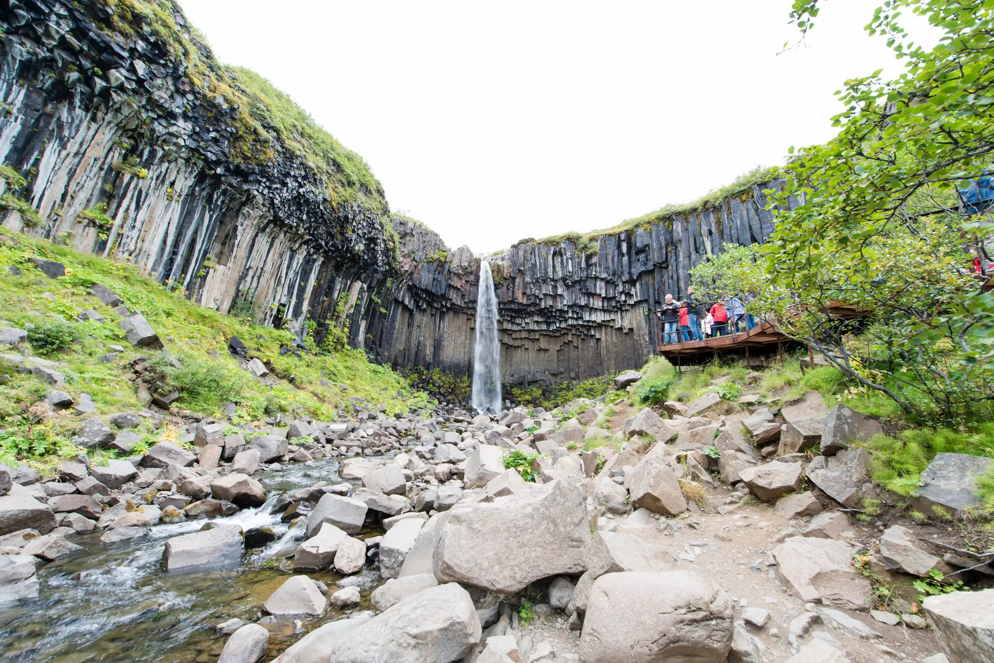 Hikers pause at a viewing platform to gaze at a waterfall cascading down among large basalt rocks.