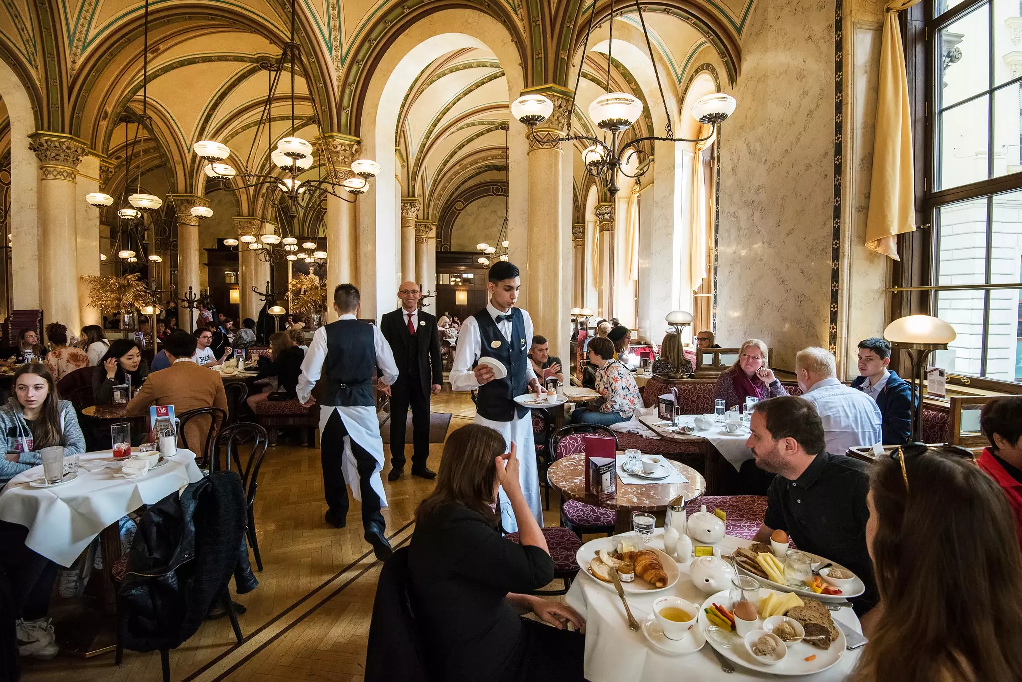 Waiters in bowties and vests serve diners seated at tables in the elaborate dining room of a restaurant.