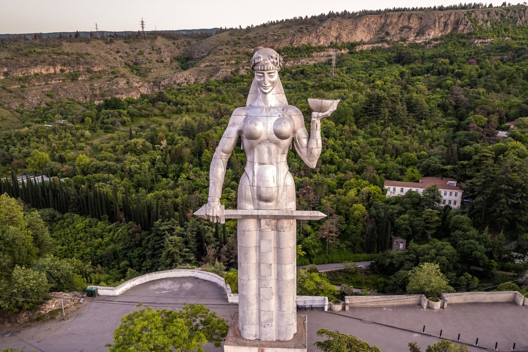 Tall silver statue of a woman seen at eye level, a green wooded hillside and a curved viewing terrace below