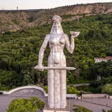 Tall silver statue of a woman seen at eye level, a green wooded hillside and a curved viewing terrace below