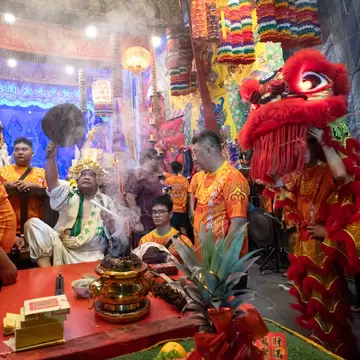 People in orange shirts surround a smoking gold urn while a man in white with a gold crown fans the smoke a chinese dragon puppet is to the right.