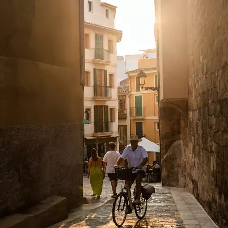 A man cycles through a narrow sunlit street in the old town of Palma de Mallorca.