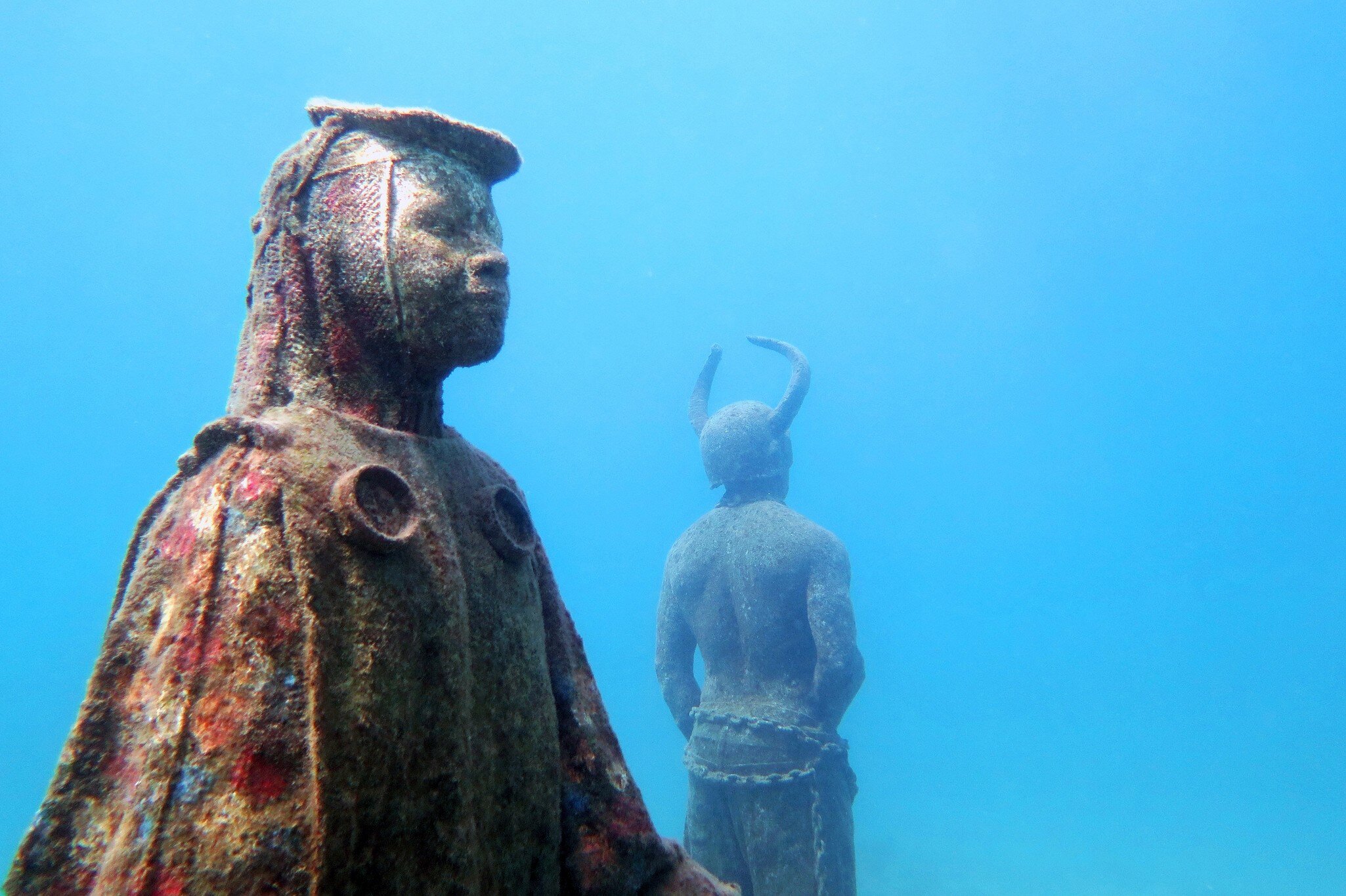 Two submerged statues coated in coral growth standing in bright blue water, one robed and facing the camera, one horned and turned away.